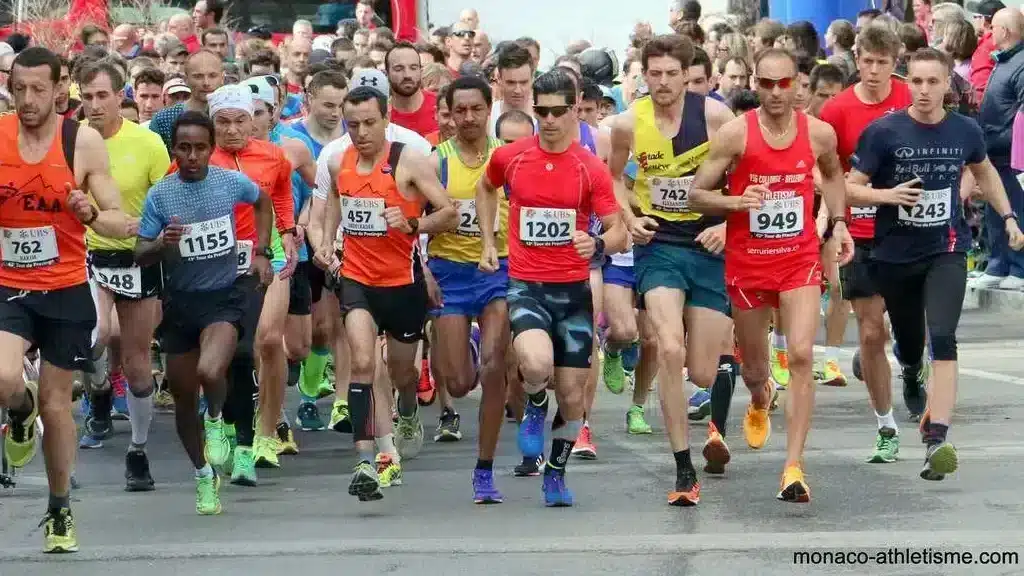 L'image montre un groupe de coureurs participant à une course à pied. Il semble être le départ de l'événement, car les coureurs sont proches les uns des autres, indiquant qu'ils viennent juste de commencer à courir. Les participants sont majoritairement des hommes, vêtus de tenues de sport variées, avec des numéros de dossard visibles sur leur torse. Les couleurs des tenues sont diverses, allant du rouge au bleu, avec des touches de jaune et d'autres couleurs vives. Certains coureurs portent des chaussures de course aux couleurs néon, ce qui est typique des équipements de haute visibilité utilisés dans les compétitions de course à pied. L'arrière-plan montre un public le long de la route, ainsi que des véhicules, indiquant que la course se déroule dans une zone urbaine ou périurbaine.