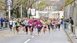 Des coureurs concentrés au début de la Course de l'Escalade à Genève, avec des spectateurs bordant la route et une affiche d'art ancestral du Gabon en arrière-plan, capturant l'énergie et le multiculturalisme de la ville