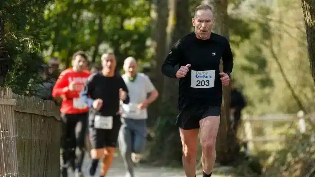 Un groupe de coureurs s'engage avec ardeur dans une course pédestre, le "Derby des Bois", comme l'indique le dossard du participant en premier plan. Ils sont encadrés par des spectateurs et évoluent dans un environnement boisé, ce qui donne une atmosphère sereine et naturelle à l'événement. L'intensité de leur effort est visible, chacun concentré sur sa performance.