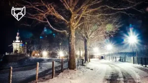 Une nuit hivernale paisible se dépeint ici, avec un chemin enneigé menant à une chapelle éclairée, tandis que les lampadaires répandent une lueur chaleureuse sous le ciel nocturne et les arbres dénudés se dressent en sentinelles silencieuses.